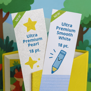 Two children’s bookmarks displayed inside a colorful kids’ book, highlighting Ultra Premium Pearl (18 pt.) with a shimmery pearl texture and Ultra Premium Smooth White (18 pt.) with a matte writable finish.